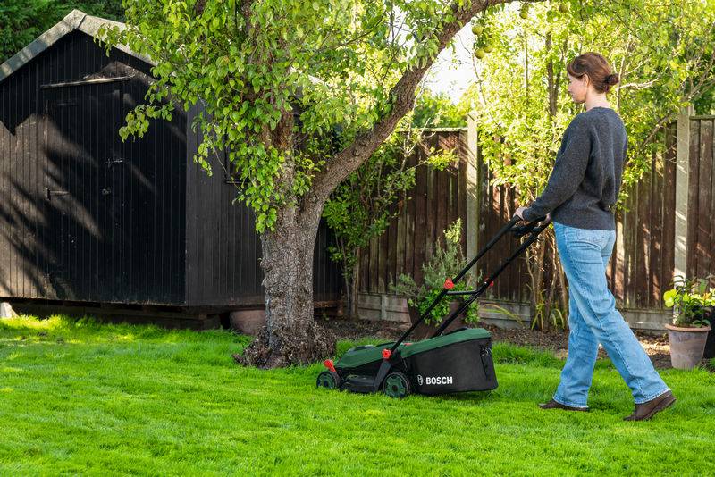 Eine Person mäht den Rasen mit einem Bosch-Rasenmäher neben einem Baum, in der Nähe eines schwarzen Schuppens und eines Holzzauns in einer Gartenumgebung.