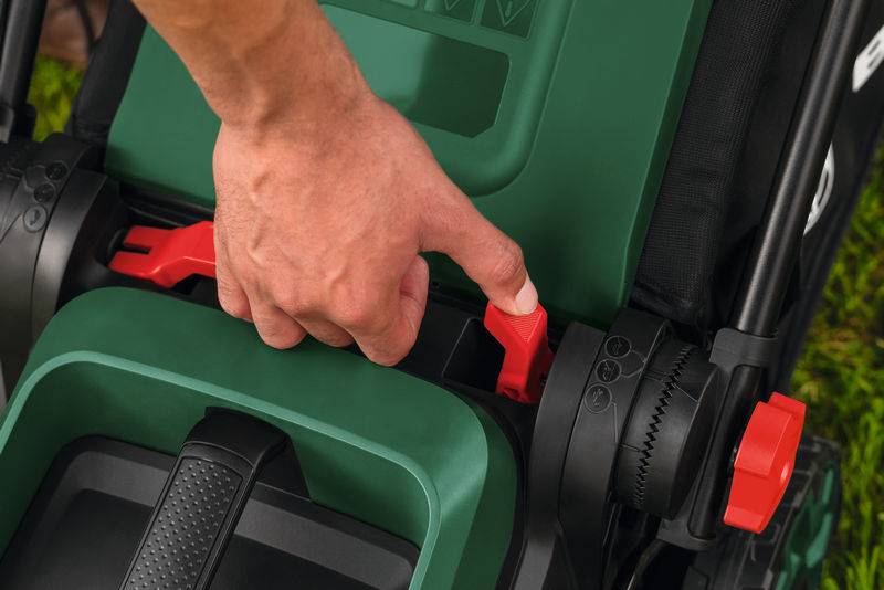 „Eine Hand verstellt die Höheneinstellung eines grünen Rasenmähers mit einem roten Hebel, was auf Benutzerfreundlichkeit und ergonomische Designmerkmale hindeutet.