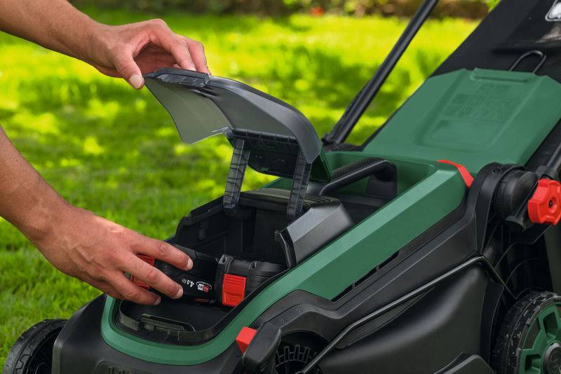 Eine Person setzt eine Batterie in einen kabellosen Rasenmäher in einer Gartenumgebung ein und zeigt dabei den Prozess der Verwendung eines elektrischen Gartengeräts.