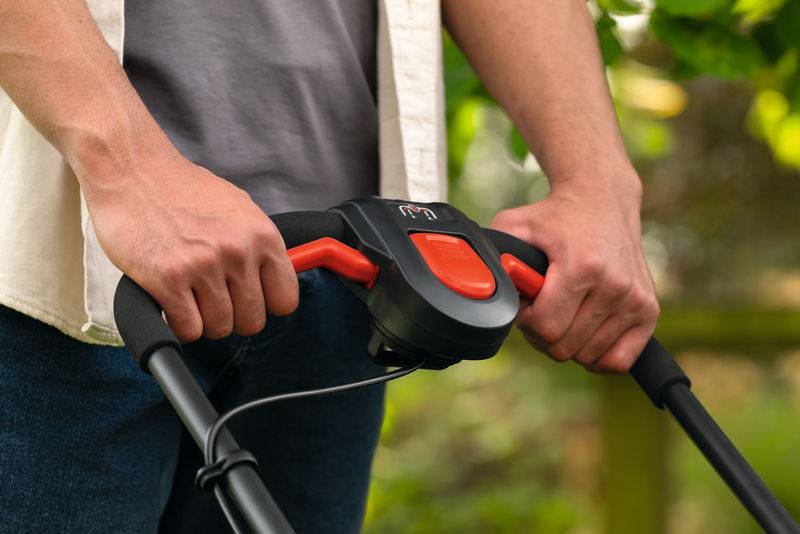 Eine Person erfasst den Griff eines Rasenmähers und konzentriert sich dabei auf die Bedienelemente. Im Hintergrund ist üppiges Grün zu sehen, das auf eine Außenumgebung hindeutet.