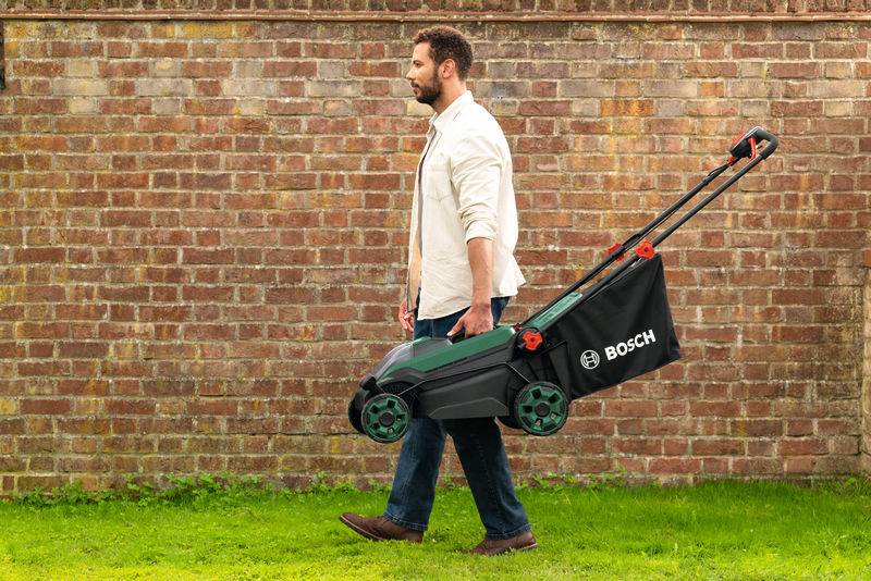 Ein Mann geht auf Gras, während er einen zusammengeklappten Rasenmäher mit einem „Bosch