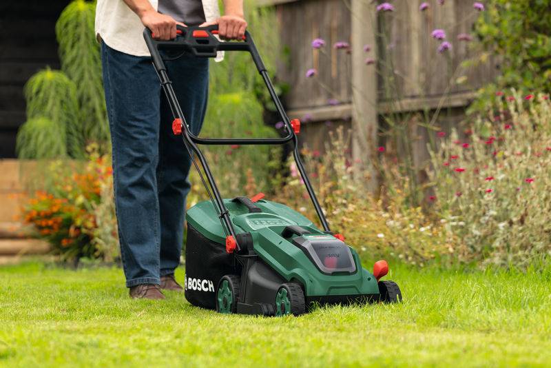 Eine Person mäht einen Rasen mit einem grünen Rasenmäher in einer Gartenumgebung. Blumen blühen im Hintergrund in der Nähe eines Holzzauns.