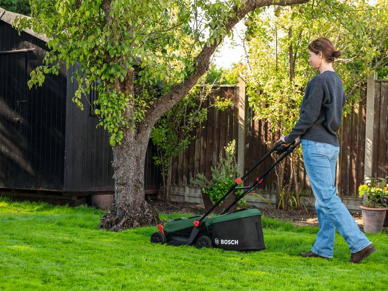 Eine Person, die mit einem Bosch-Rasenmäher einen Rasen in einem Garten mäht, in der Nähe eines Baumes und eines Holzschuppens, an einem sonnigen Tag.