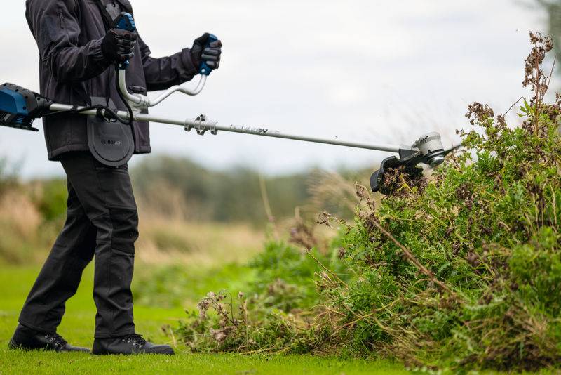 Eine Person, die schwarze Schutzausrüstung trägt, schneidet überwucherte Büsche mit einem Trimmer auf einer Wiese ab und konzentriert sich auf die Beseitigung von Vegetation.
