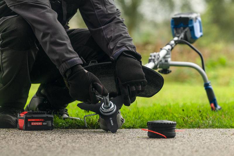 Eine Person, die den Aufsatz an einem Rasentrimmer wechselt, der auf Gras ruht. Ein Akku und Trimmerteile sind auf dem Boden sichtbar.