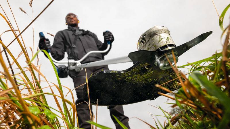 Eine Person bedient einen Freischneider und schneidet hohes Gras. Das Bild konzentriert sich auf die Klingen des Freischneiders und hebt seine Kraft und Effizienz hervor.