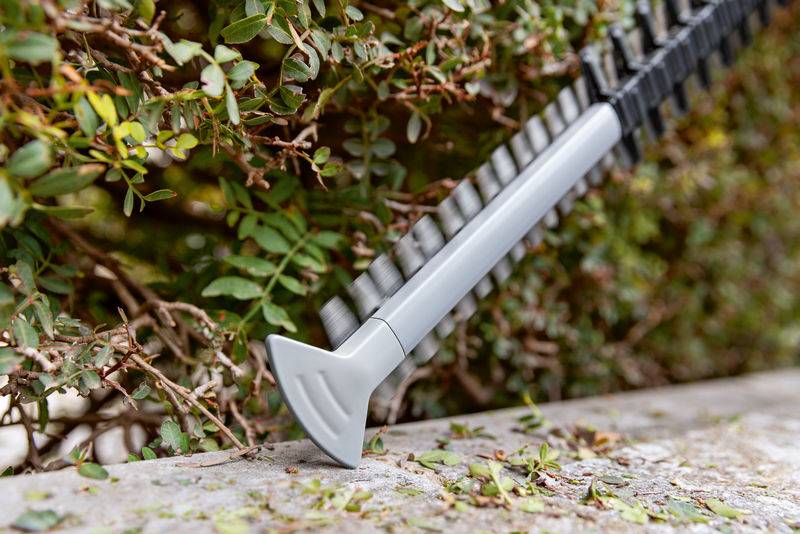 Eine Heckenschere ruht auf einer Steinkante, umgeben von grünem Laub. Die Klinge der Schere zeigt nach unten, in einem Winkel in die Hecke.