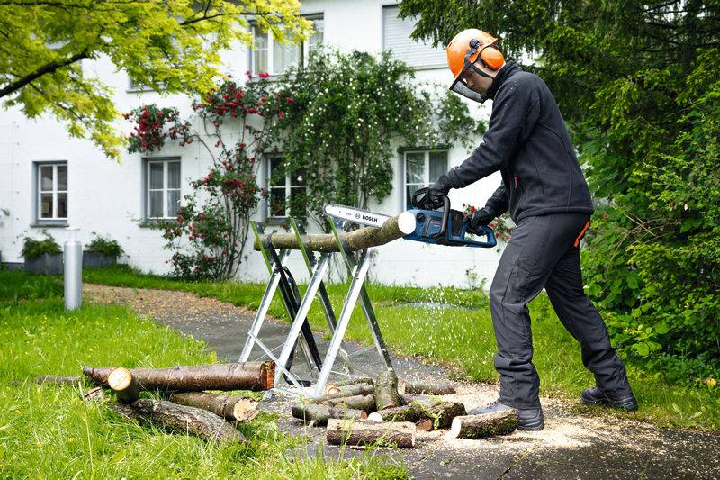 Eine Person in Schutzausrüstung benutzt eine Kettensäge, um einen Baumstamm auf einem Metallständer im Garten zu schneiden, mit einem weißen Haus und Grünflächen im Hintergrund.