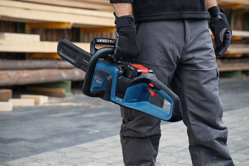 Eine Person hält eine blau-schwarze Kettensäge. Sie trägt graue Hosen und Handschuhe. Holzbretter und Baumstämme sind im Hintergrund sichtbar.