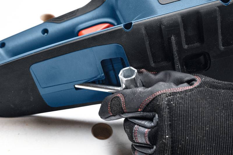 Eine Hand mit einem schwarzen Handschuh verwendet einen Schraubenschlüssel, um ein Teil an einem blau-schwarzen Elektrowerkzeug einzustellen und betont damit Wartung oder Reparatur.