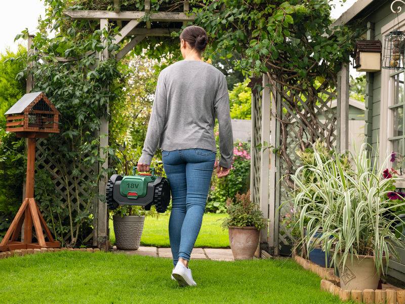 Eine Person geht durch einen Garten und hält einen grünen Roboter-Rasenmäher, umgeben von Grünflächen und einem Vogelhaus.
