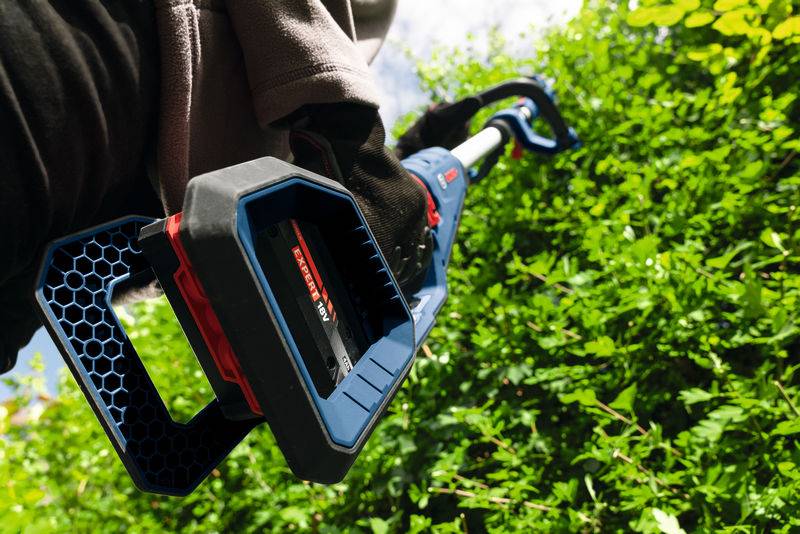 Eine Person schneidet eine Hecke mit einer schnurlosen elektrischen Heckenschere, wobei sie diese diagonal nach oben in Richtung dichten grünen Laubwerks unter einem klaren Himmel hält.