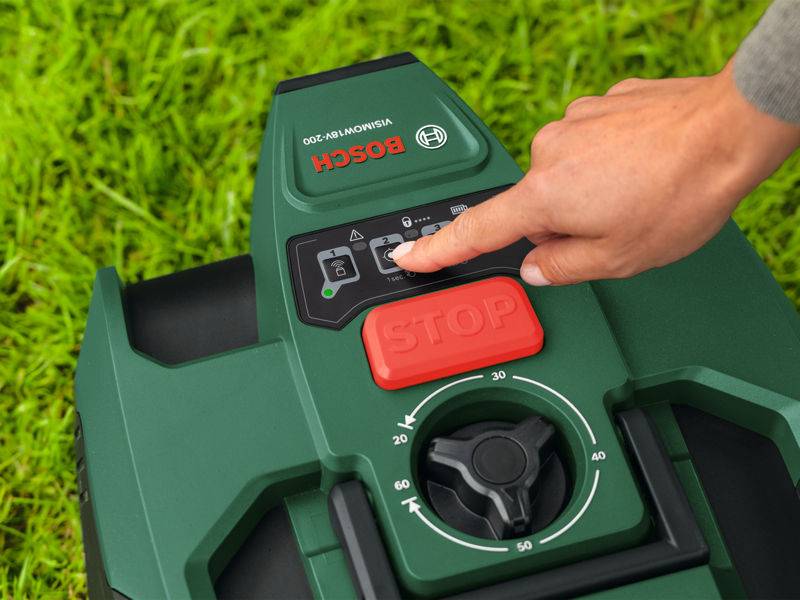 „Die Hand einer Person drückt einen Knopf auf einem grünen autonomen Rasenmäher von Bosch, der auf Gras steht, wobei der Fokus auf der Bedienplatte und der roten STOPP-Taste liegt.