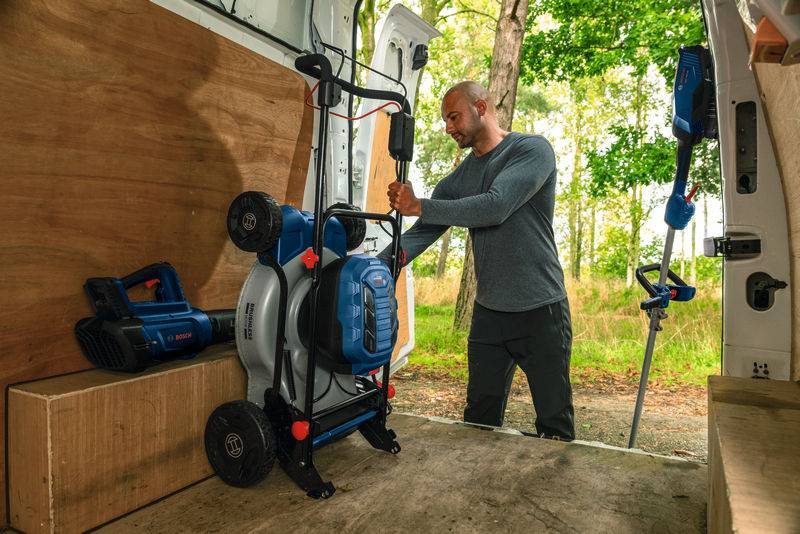 Eine Person in einem Transporter sichert einen blauen Hochdruckreiniger und Batteriegeräte. Bäume sind durch die offene Tür sichtbar, was auf Vorbereitung für Außenarbeiten hindeutet.