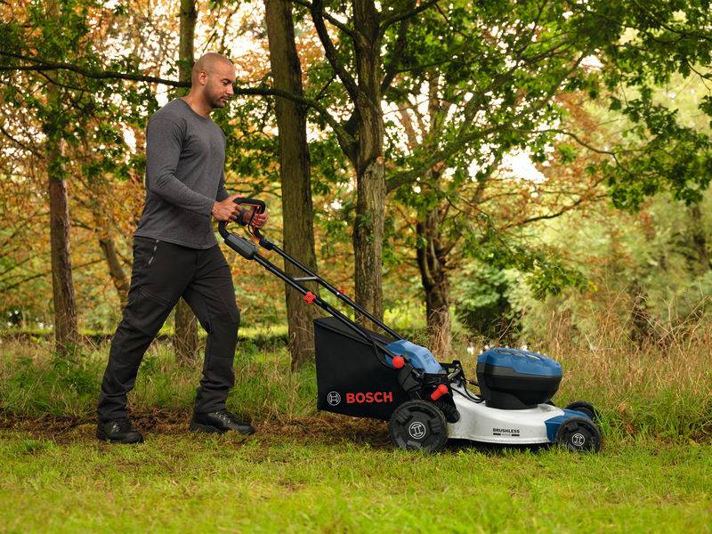 Ein Mann mäht einen grasbewachsenen Rasen mit einem kabellosen Bosch-Rasenmäher in einer Parkanlage, umgeben von Bäumen mit Herbstlaub.