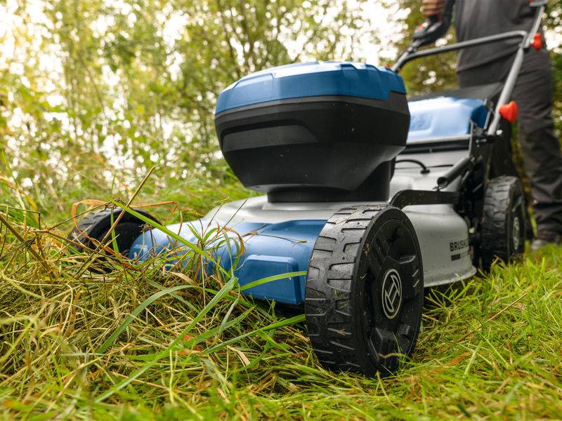 Eine Person schiebt einen blau-schwarzen Rasenmäher, der wildes Gras in einem Garten schneidet, umgeben von grünen Büschen und Bäumen.