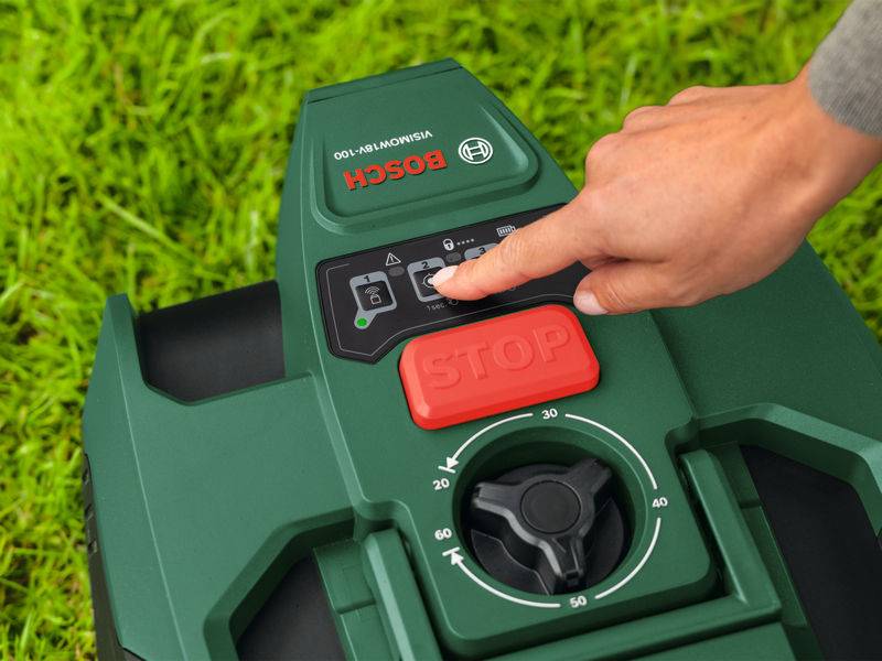 Eine Hand, die die Starttaste auf einem Bosch-Roboterrasenmäher drückt, der auf Gras steht, mit sichtbarem Bedienfeld und Notausschalter.