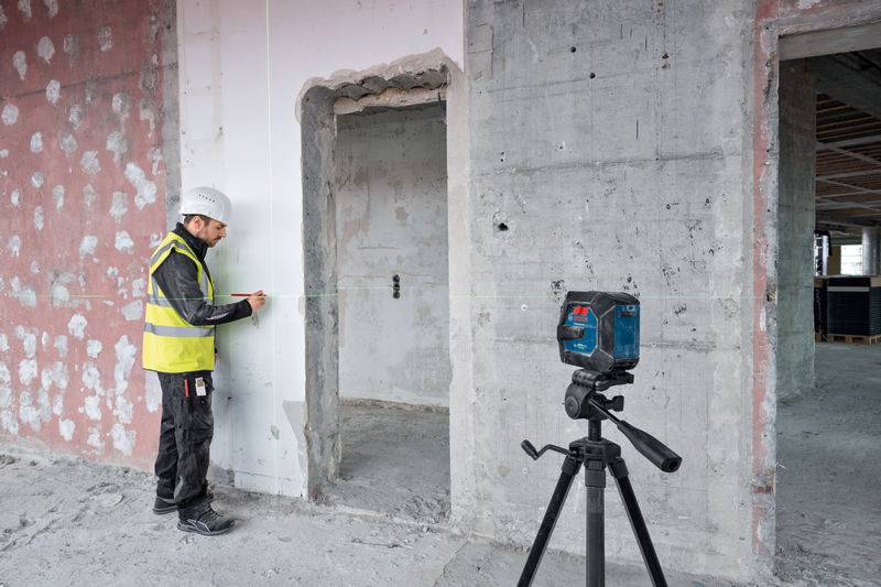 Ein Bauarbeiter in einer gelben Weste verwendet ein Laser-Nivelliergerät, um eine Bleistiftmarkierung an einer Wand in einem sich im Bau befindlichen Gebäude zu führen.