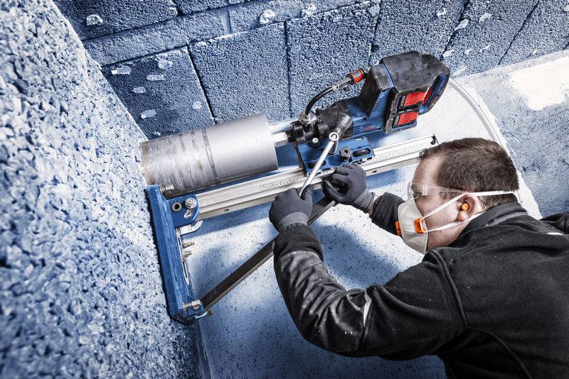 Ein Arbeiter in Schutzausrüstung bedient einen blauen Kernbohrer und bohrt ein Loch in eine Betonwand. Die Szene betont Industriesicherheit.