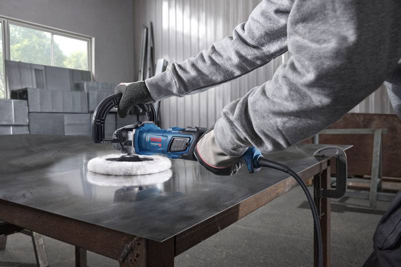 Eine Person nutzt einen elektrischen Schleifer auf einem Metalltisch in einer Industrieumgebung, trägt Schutzhandschuhe und ein Sweatshirt.