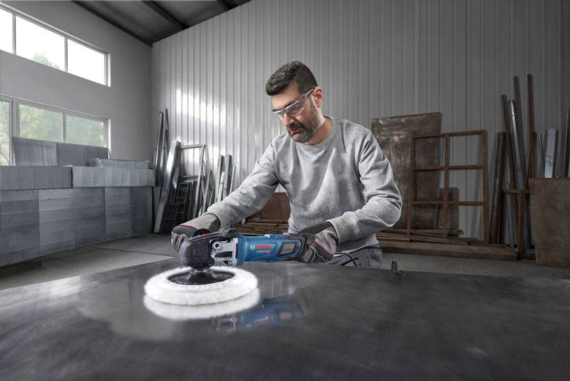 Ein Mann, der eine Schutzbrille und Handschuhe trägt, verwendet ein Elektrowerkzeug, um eine große Metalloberfläche in einer Industriewerkstatt zu polieren.