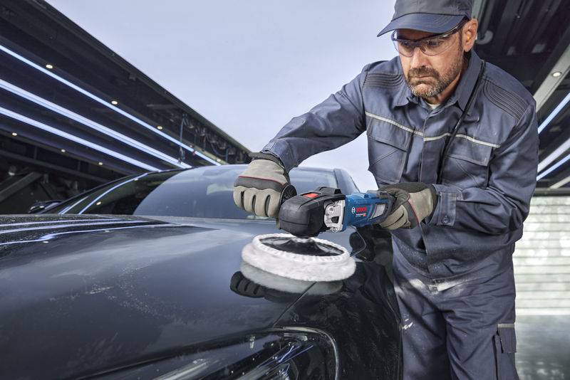 Ein Mann, der Schutzausrüstung trägt, poliert die Motorhaube eines Autos mit einem Elektrowerkzeug in einer Werkstattumgebung und konzentriert sich intensiv auf die Aufgabe.