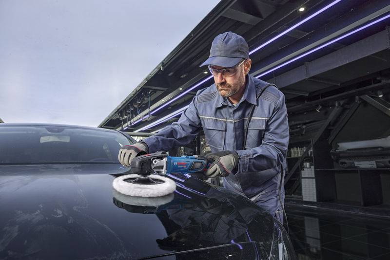 Eine Person in blauer Uniform und Mütze verwendet einen elektrischen Poliermaschine, um die Motorhaube eines Autos in einer Garage mit moderner Beleuchtung zu polieren.