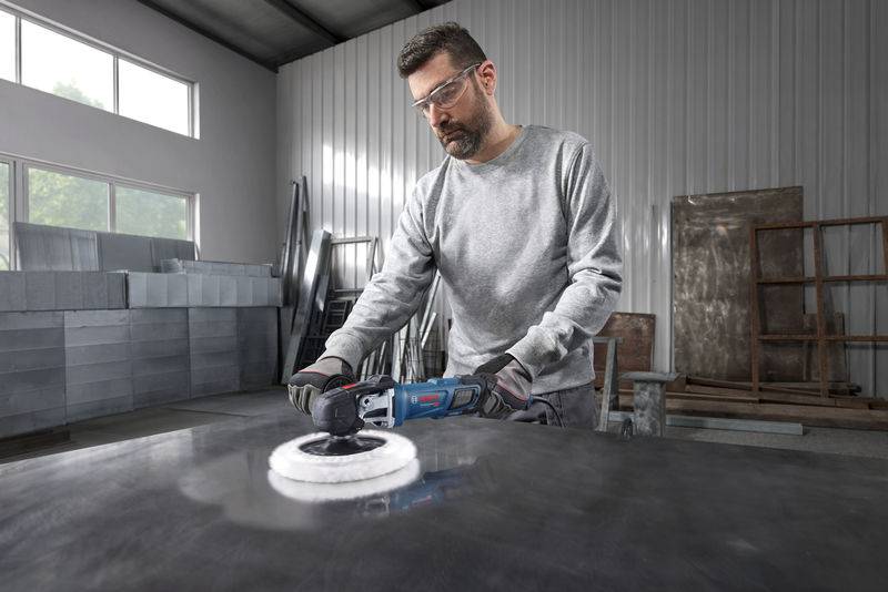 Eine Person in einer Werkstatt verwendet ein elektrisches Poliergerät auf einer Metalloberfläche, trägt eine Schutzbrille und Handschuhe, mit Industrieausrüstung in der Umgebung.