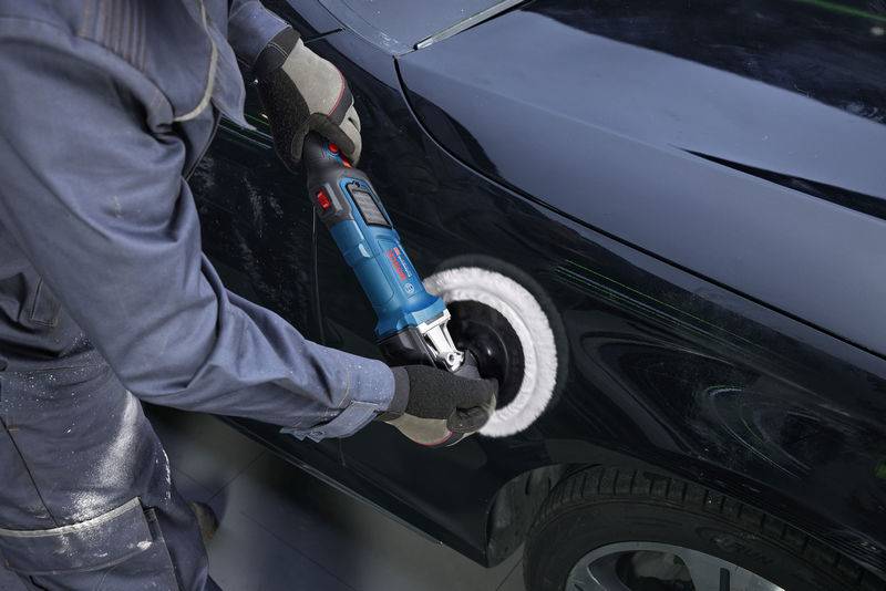 Eine Person benutzt einen blau-schwarzen elektrischen Poliermaschine, um ein glänzendes dunkles Auto zu polieren, während sie Schutzkleidung und Handschuhe trägt.
