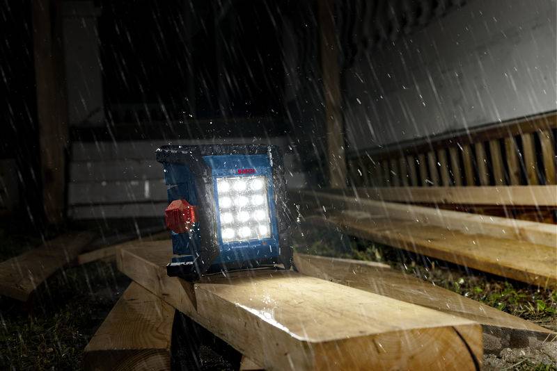 Ein tragbares LED-Arbeitslicht beleuchtet eine Baustelle nachts bei starkem Regen und hebt Holzbretter und teilweise errichtete Strukturen hervor.