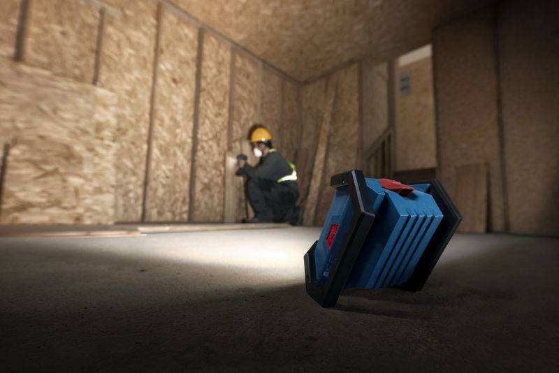 Ein Bauarbeiter mit Schutzhelm und Weste macht ein Foto in einem unfertigen Holzrahmenzimmer, während eine blaue tragbare Leuchte im Vordergrund steht.