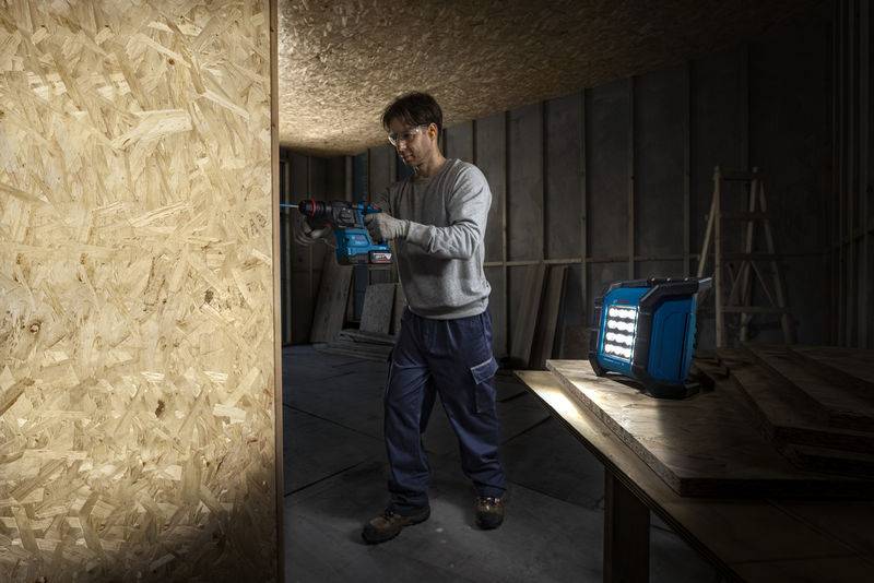 Eine Person in einem schwach beleuchteten Baubereich, die eine Elektrobohrmaschine auf einer Holzplatte verwendet. Eine helle Arbeitsleuchte beleuchtet den Arbeitsbereich.