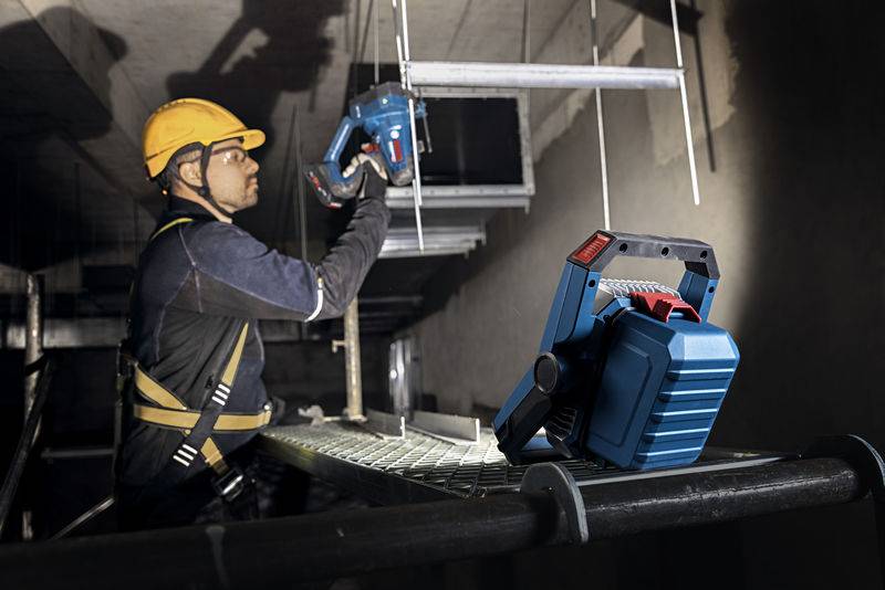 Ein Arbeiter, der einen Helm und ein Sicherheitsgeschirr trägt, verwendet ein kabelloses Elektrowerkzeug an einem Metallrahmen in Innenräumen, während ein anderes Werkzeug in der Nähe liegt.