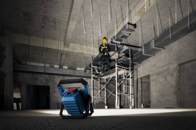 Eine Person auf Gerüsten in einem großen, schwach beleuchteten Industrieraum, mit Fokus auf ein tragbares blaues Arbeitslicht im Vordergrund.