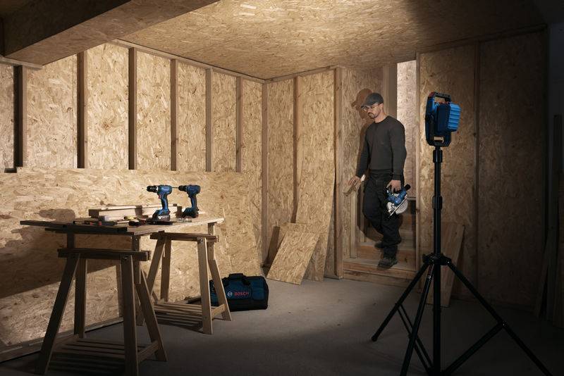 Eine Person in einer Kappe und Brille betritt einen teilweise fertiggestellten Holzraum und hält eine Säge in der Hand. Werkzeuge und ein Lichtständer befinden sich in der Nähe.
