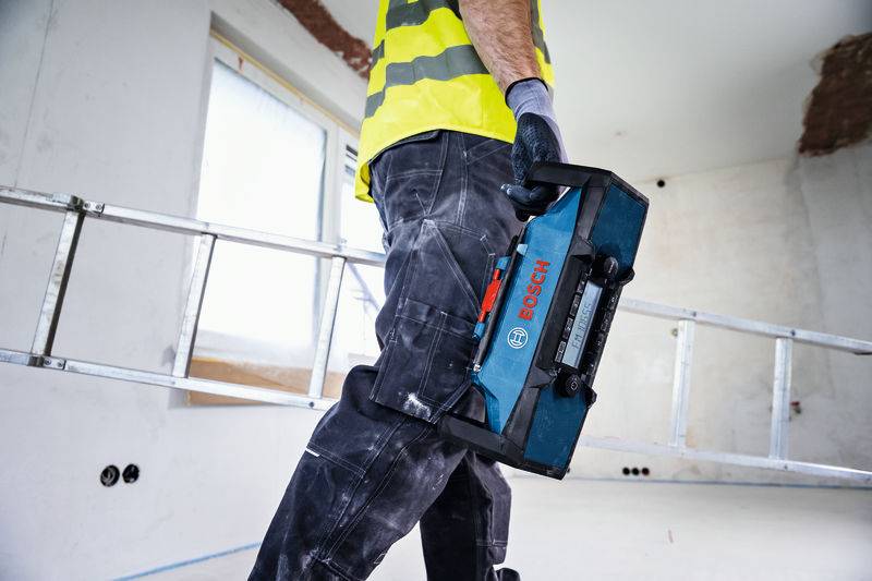Ein Arbeiter in Baukleidung trägt einen Bosch-Werkzeugkoffer und geht durch einen unfertigen Raum mit einer Leiter und Flicken an den Wänden.