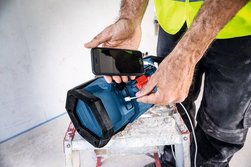 Eine Person in Baukleidung verbindet ein Smartphone mit einem blauen Werkzeug mit einem roten Stecker, möglicherweise zum Aufladen oder Verbinden von Geräten.