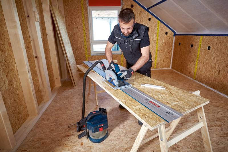 Eine Person in Schutzausrüstung verwendet eine Kreissäge, um Holz in einem teilweise konstruierten Holzzimmer zu schneiden.