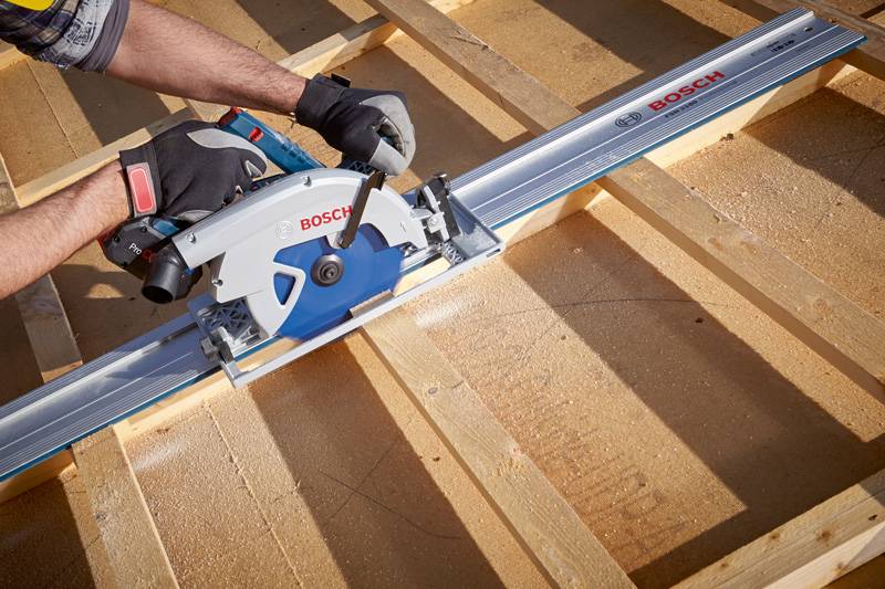 Eine Person nutzt eine Kreissäge mit einer Bosch-Führungsschiene, um Holz auf einer Baustelle zu schneiden. Die Säge hat ein blaues Blatt und der Bediener trägt Handschuhe.