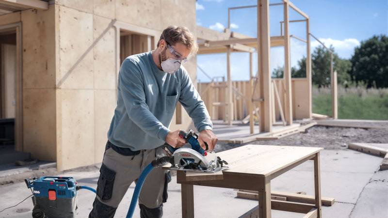 Eine Person in Schutzausrüstung verwendet eine Kreissäge auf Holz auf einer Baustelle mit teilweise errichteten Holzstrukturen im Hintergrund.