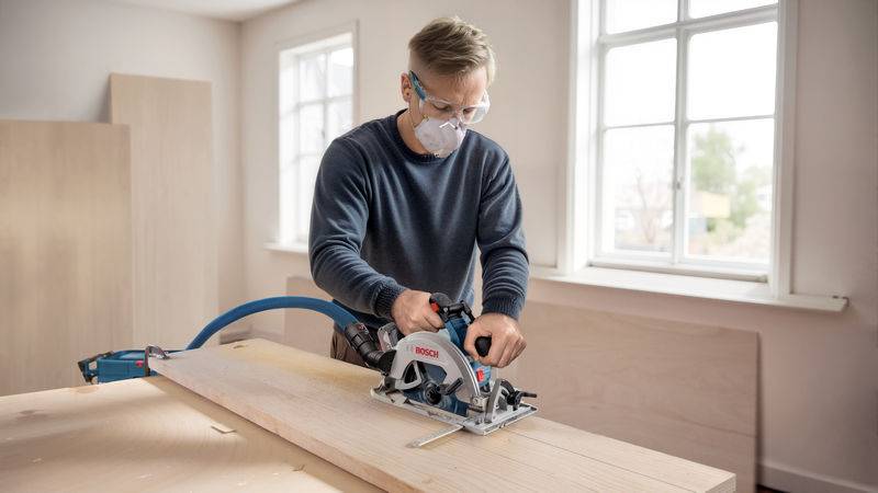 Eine Person, die eine Schutzbrille und eine Maske trägt, benutzt eine Kreissäge, um ein Holzbrett in einer gut beleuchteten Werkstatt zu schneiden.