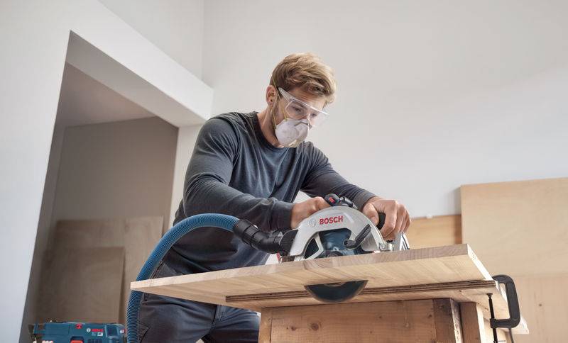 Eine Person in Schutzausrüstung bedient eine Bosch-Elektrosäge und schneidet ein Holzbrett auf einem Tisch in einer Werkstatt.