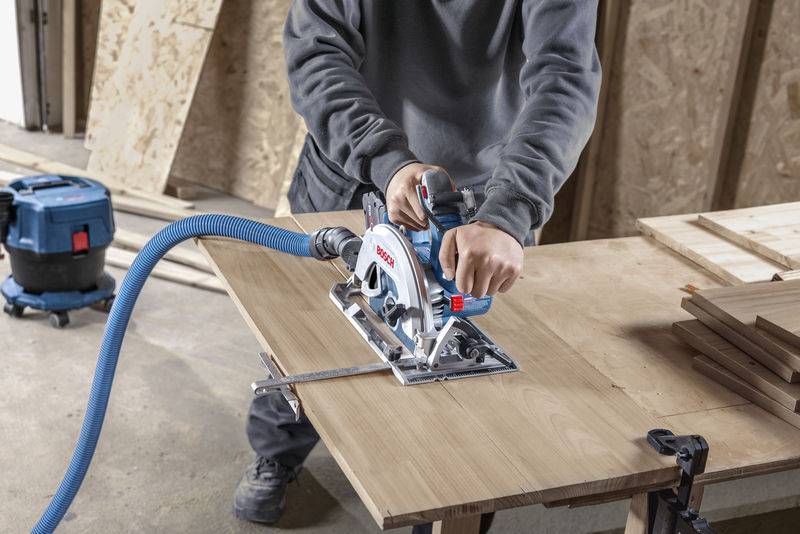 Eine Person benutzt eine Kreissäge, um ein Holzbrett auf einer Werkbank zu schneiden, mit einem angeschlossenen Staubsaugerschlauch zur Staubabsaugung.