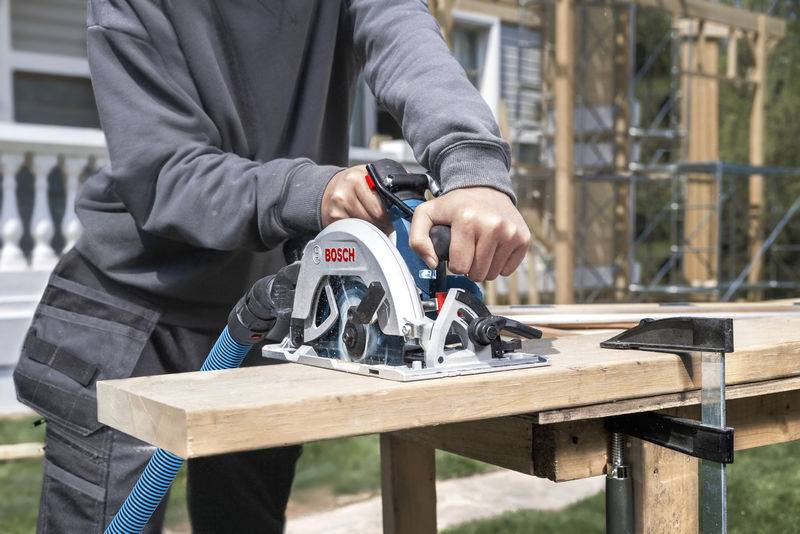 Person, die eine Bosch-Kreissäge verwendet, um Holz auf einer Werkbank zu schneiden, trägt einen grauen Pullover, mit Baugerüsten im Hintergrund.
