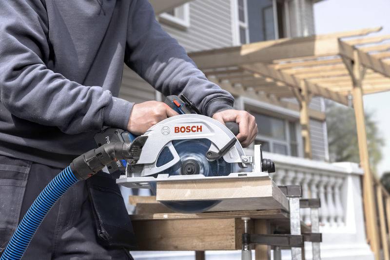 Eine Person nutzt eine Bosch-Kreissäge, um draußen ein Stück Holz zu schneiden, mit einer Holzlaube und einem Haus im Hintergrund.