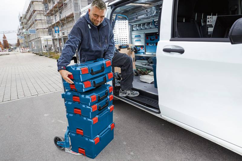 Ein Arbeiter entlädt einen Stapel blauer und roter Werkzeugkästen aus der Rückseite eines Lieferwagens, der auf einer Straße in der Nähe einer Baustelle geparkt ist.