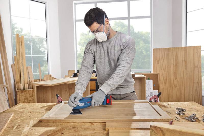 Eine Person, die eine Schutzmaske trägt, schleift eine Holzschranktür in einer Werkstatt, umgeben von Werkzeugen und Holzplatten, mit großen Fenstern im Hintergrund.