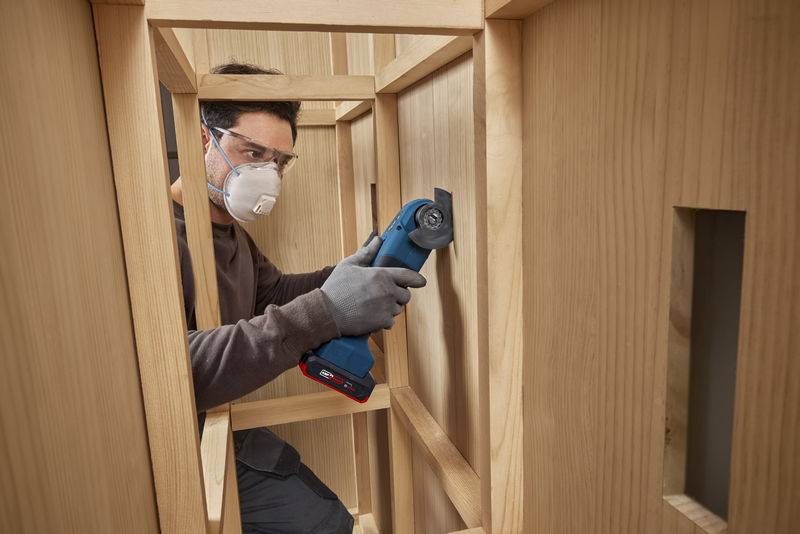 Person mit Schutzbrille und Maske, die eine elektrische Säge in einer Holzkonstruktion verwendet und dabei Konzentration und Sicherheit bei Bauarbeiten vermittelt.