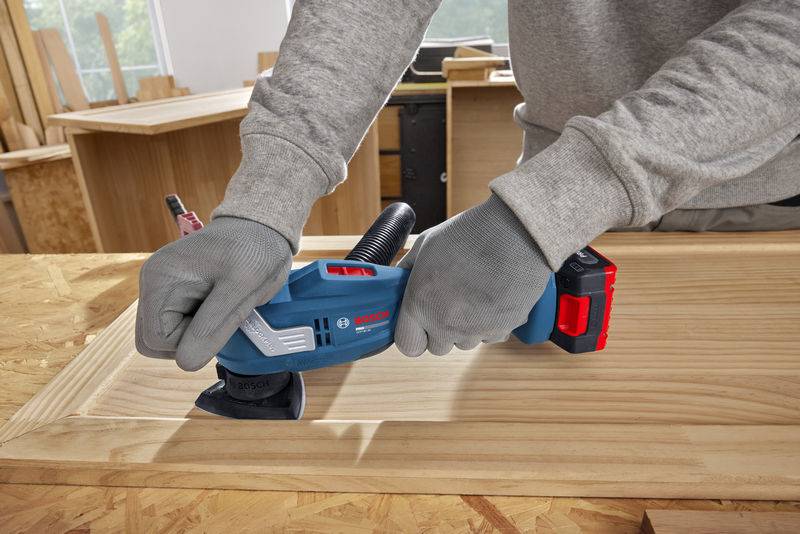 Eine Person in grauen Handschuhen schleifen eine Holzoberfläche mit einem elektrischen Elektroschleiffer von Bosch in einer Werkstatt.