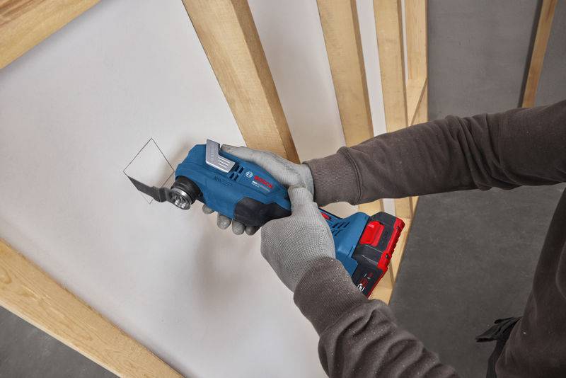 Eine Person, die ein blaues oszillierendes Elektrowerkzeug verwendet, um ein weißes Brett zu schneiden, trägt graue Handschuhe und ein langärmliges Hemd.